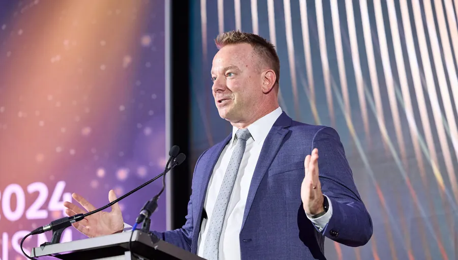 Man speaking at a podium in front of a patterned backdrop at a conference