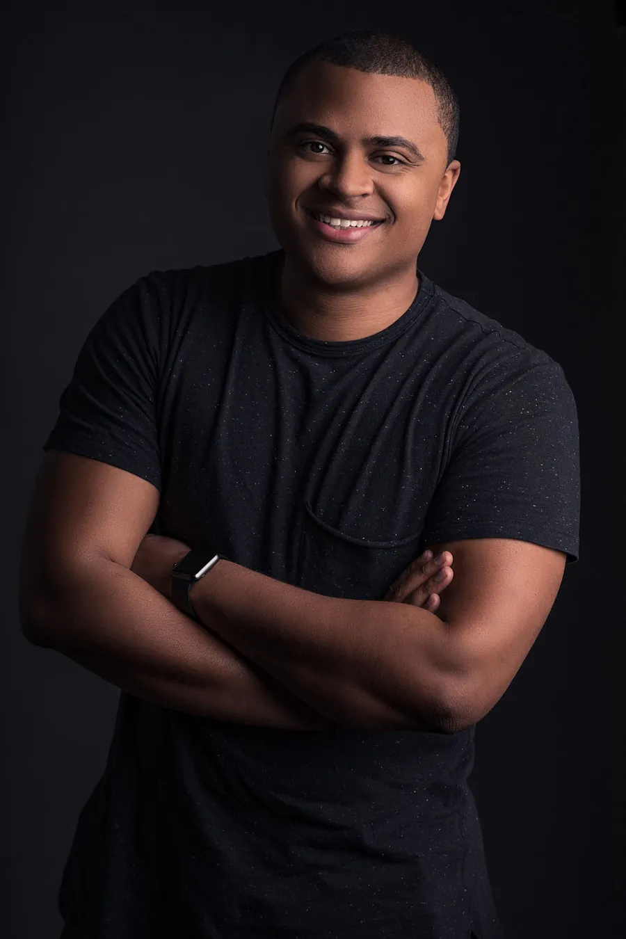 Man with crossed arms in a black t-shirt against a dark background, headshot