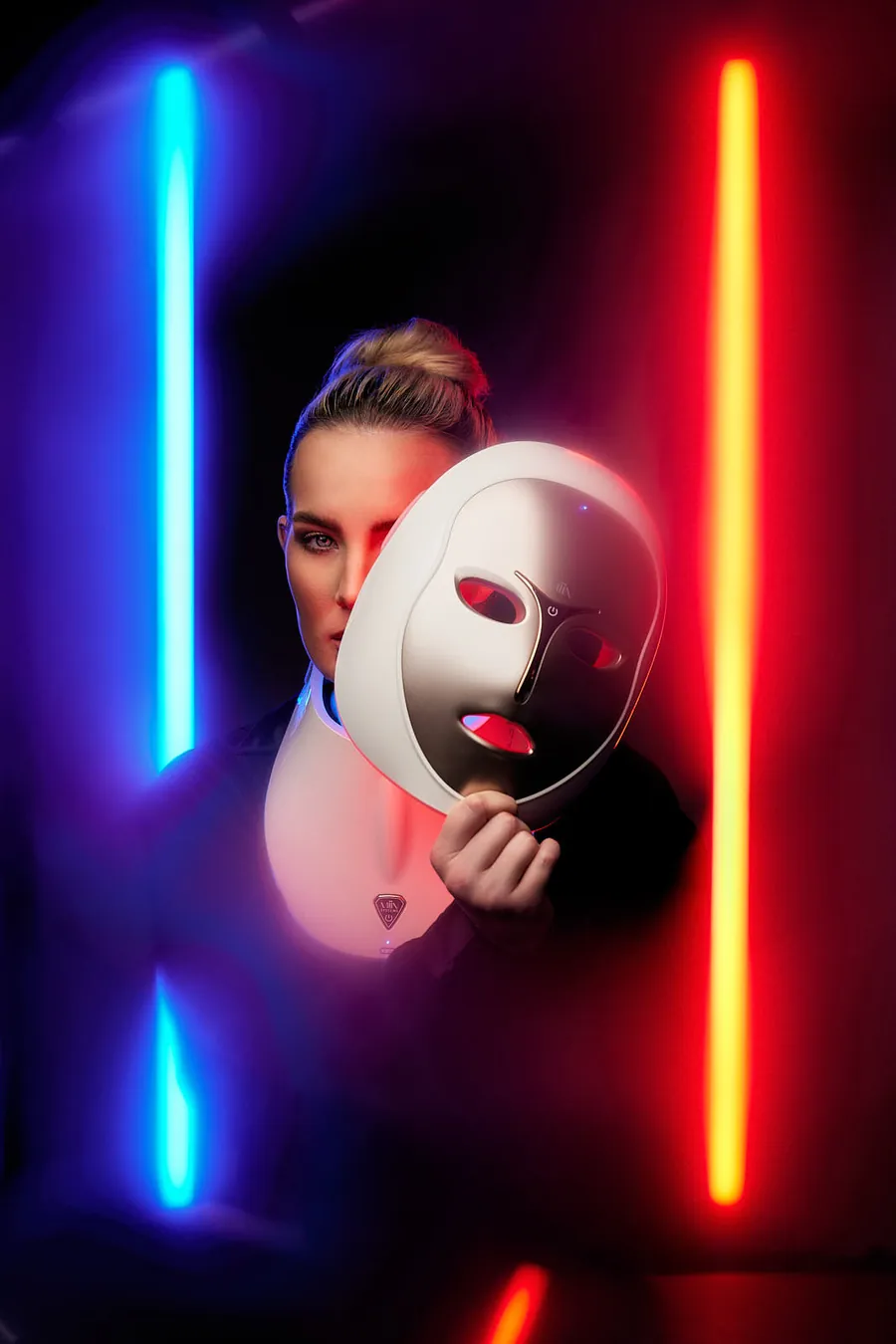 Model holding an illuminated LED light therapy mask between glowing red and blue neon lights, branding session for Artemis