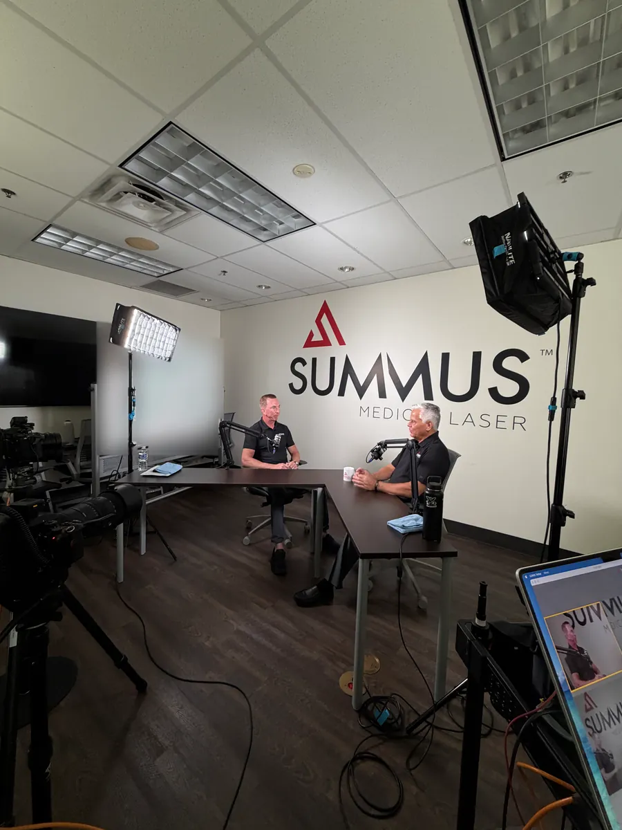 Two men recording a podcast interview at a desk in front of a Summus Medical Laser wall logo, with studio lights and cameras around the set