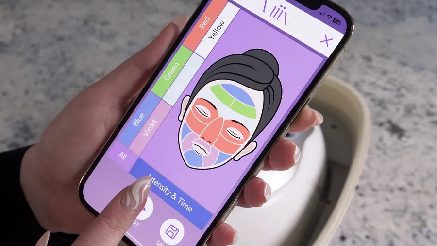 Hand holding a phone showing the MiiN LED mask app's color treatment zones, with the mask device behind it, from an Artemis product video