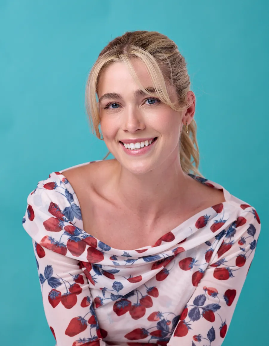 Smiling commercial actor headshot of a young woman with a blonde bun in a red and blue floral top against a teal background