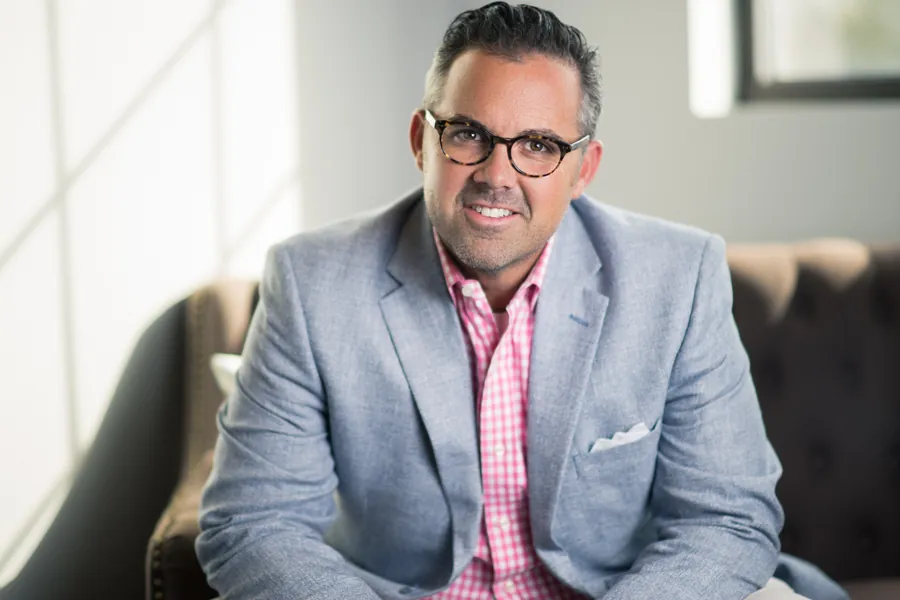Smiling man in a light gray blazer and pink checked shirt on a couch, corporate headshot