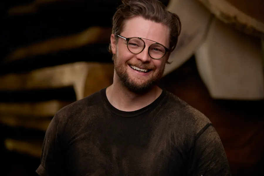 Smiling man with glasses in a dark shirt in front of wood slabs, branding portrait