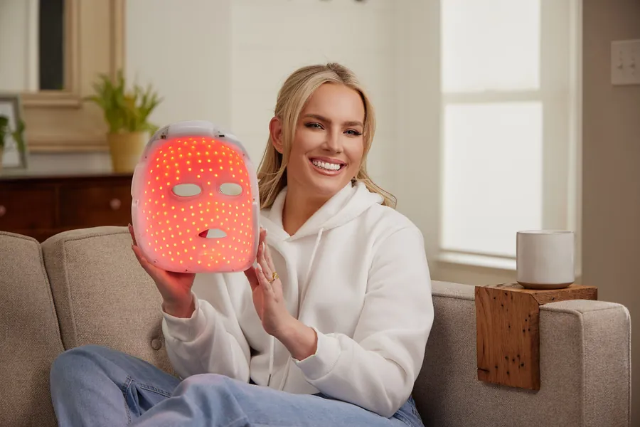 Smiling woman in a white sweatshirt holding a glowing LED mask on a couch, Artemis lifestyle photo