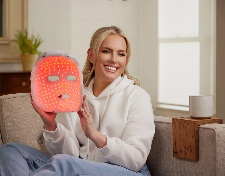 Smiling woman in a white sweatshirt holding a glowing LED mask on a couch, Artemis lifestyle photo