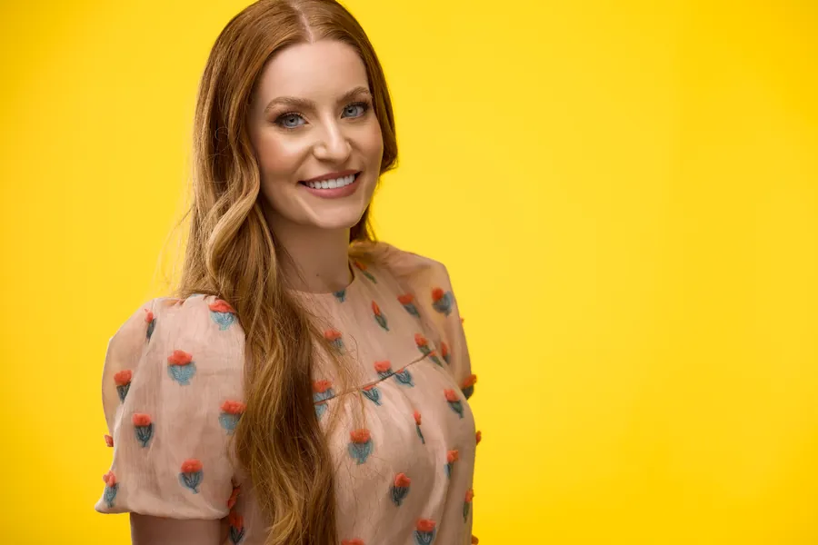 Smiling woman with red hair in a peach floral top against a yellow background, professional headshot