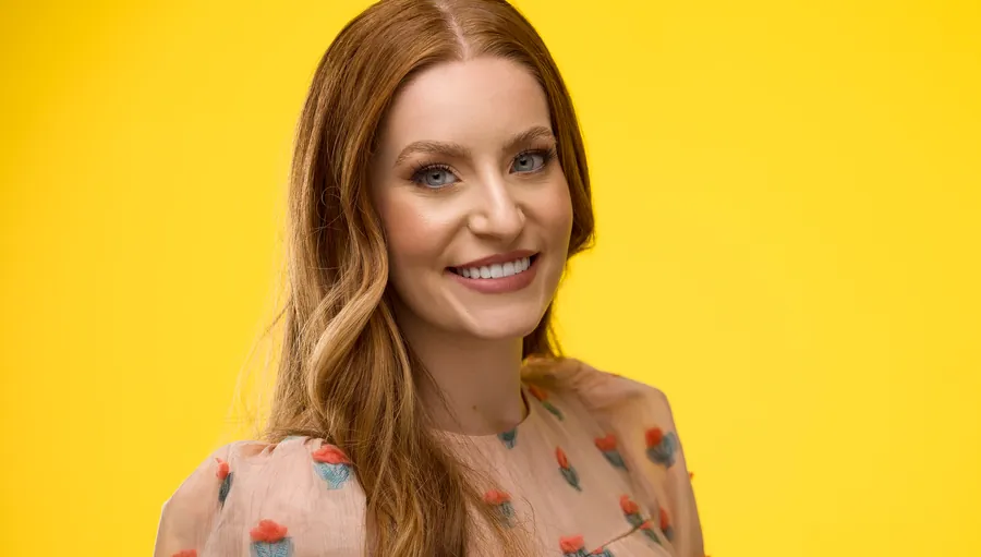 Smiling woman with red hair in a peach floral top against a yellow background, professional headshot