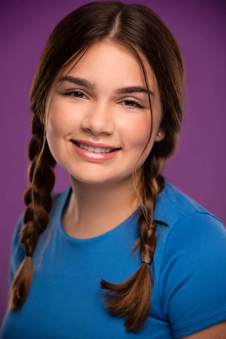 Smiling young actor headshot of a girl with two dark braids in a blue t-shirt against a purple background