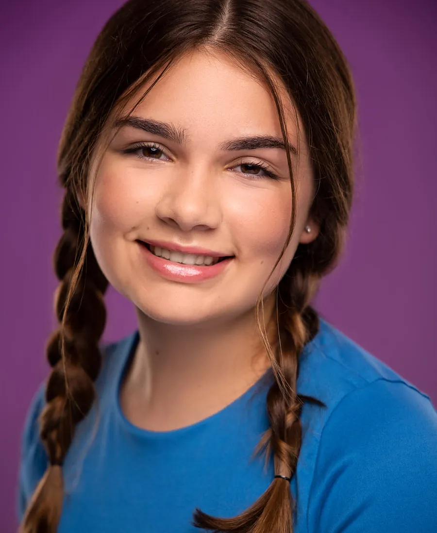 Smiling young actor headshot of a girl with two dark braids in a blue t-shirt against a purple background