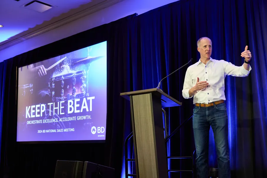 Speaker giving a thumbs up at a podium beside a large presentation screen at a general session