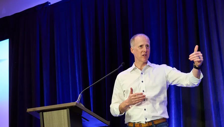Speaker giving a thumbs up at a podium beside a large presentation screen at a general session