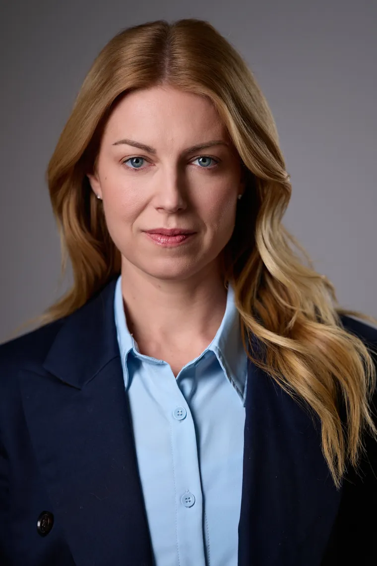 Theatrical acting headshot of a woman with long blonde hair in a navy blazer and light blue shirt against a gray background