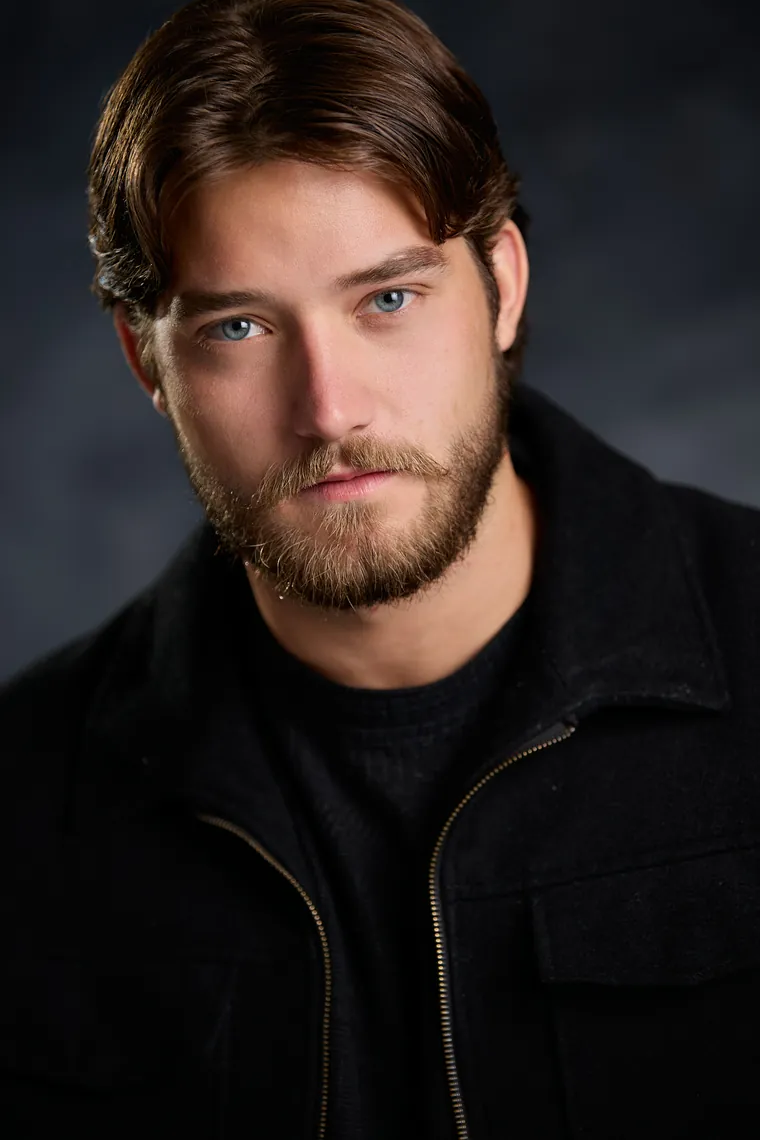 Theatrical acting headshot of a bearded man with swept brown hair in a dark jacket against a dark gray background