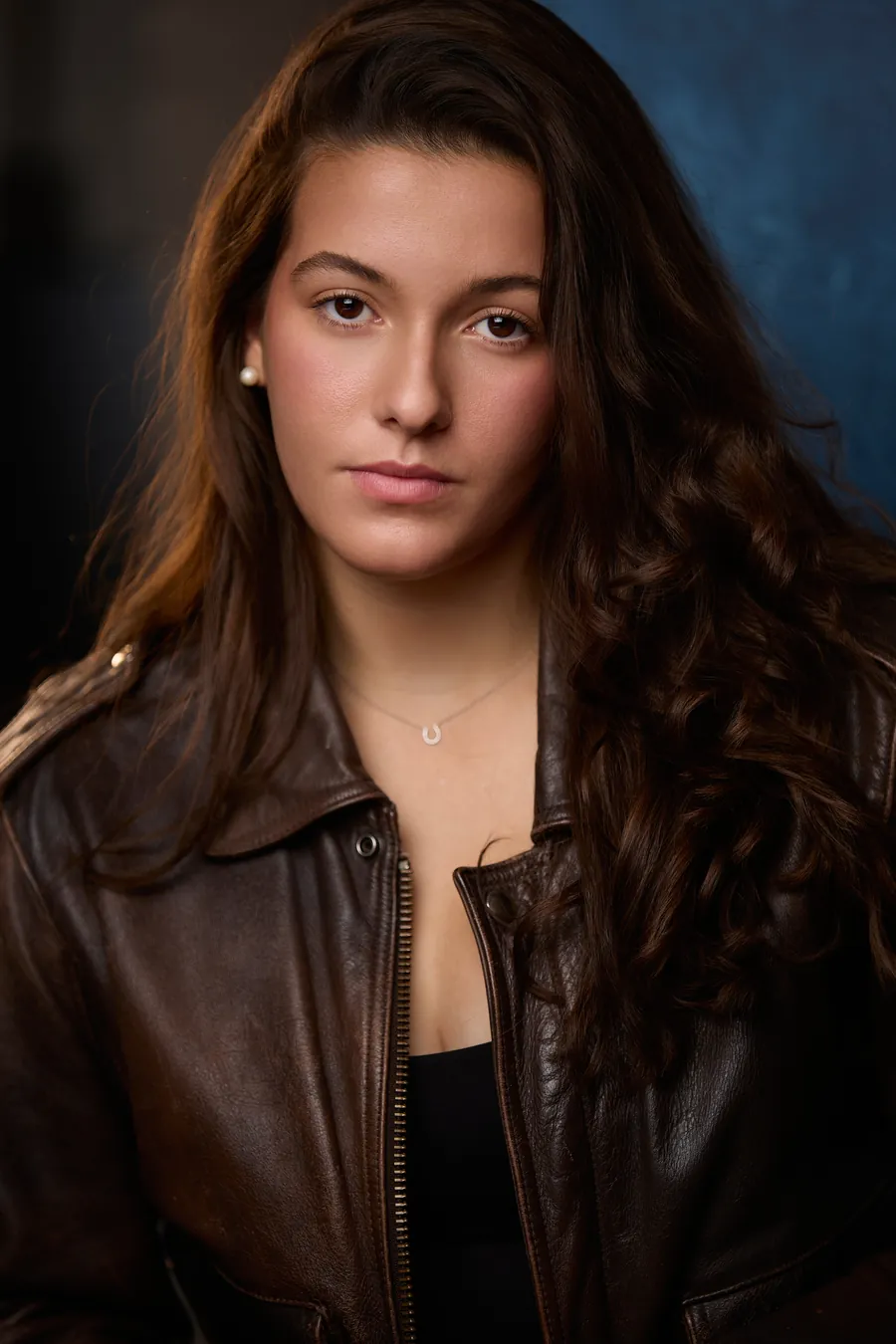 Theatrical actor headshot of a young woman with long dark wavy hair in a brown leather jacket against a dark blue background