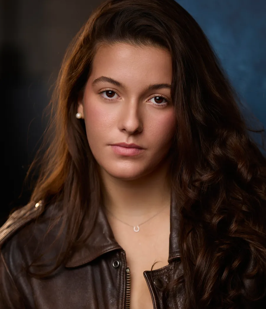 Theatrical actor headshot of a young woman with long dark wavy hair in a brown leather jacket against a dark blue background