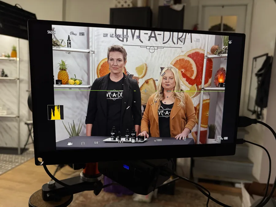 Two people on camera in front of a Give-A-Derm branded set, monitor showing the live shot