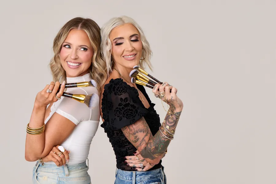 Two women back to back holding makeup brushes and smiling, High Contrast Beauty branding photo