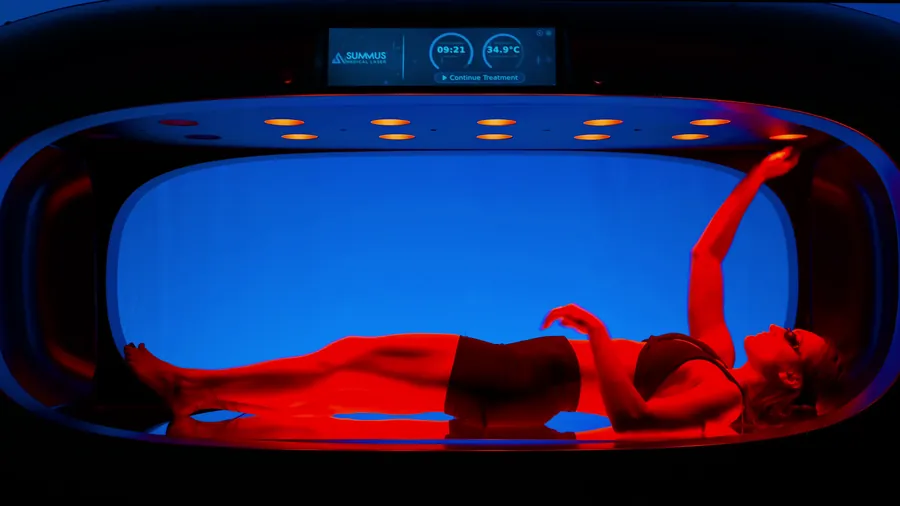 Woman lying in a Summus red light therapy pod, lit red against the glowing blue interior, Summus Pod sizzle reel — click to play