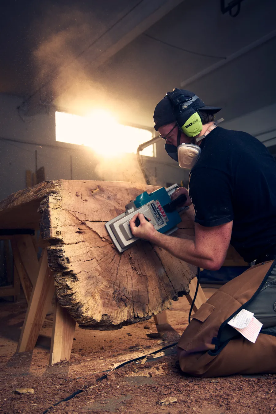 Woodworker in a mask cutting a live-edge slab lit by a bright light, branding photography for Sawdust & Whiskey